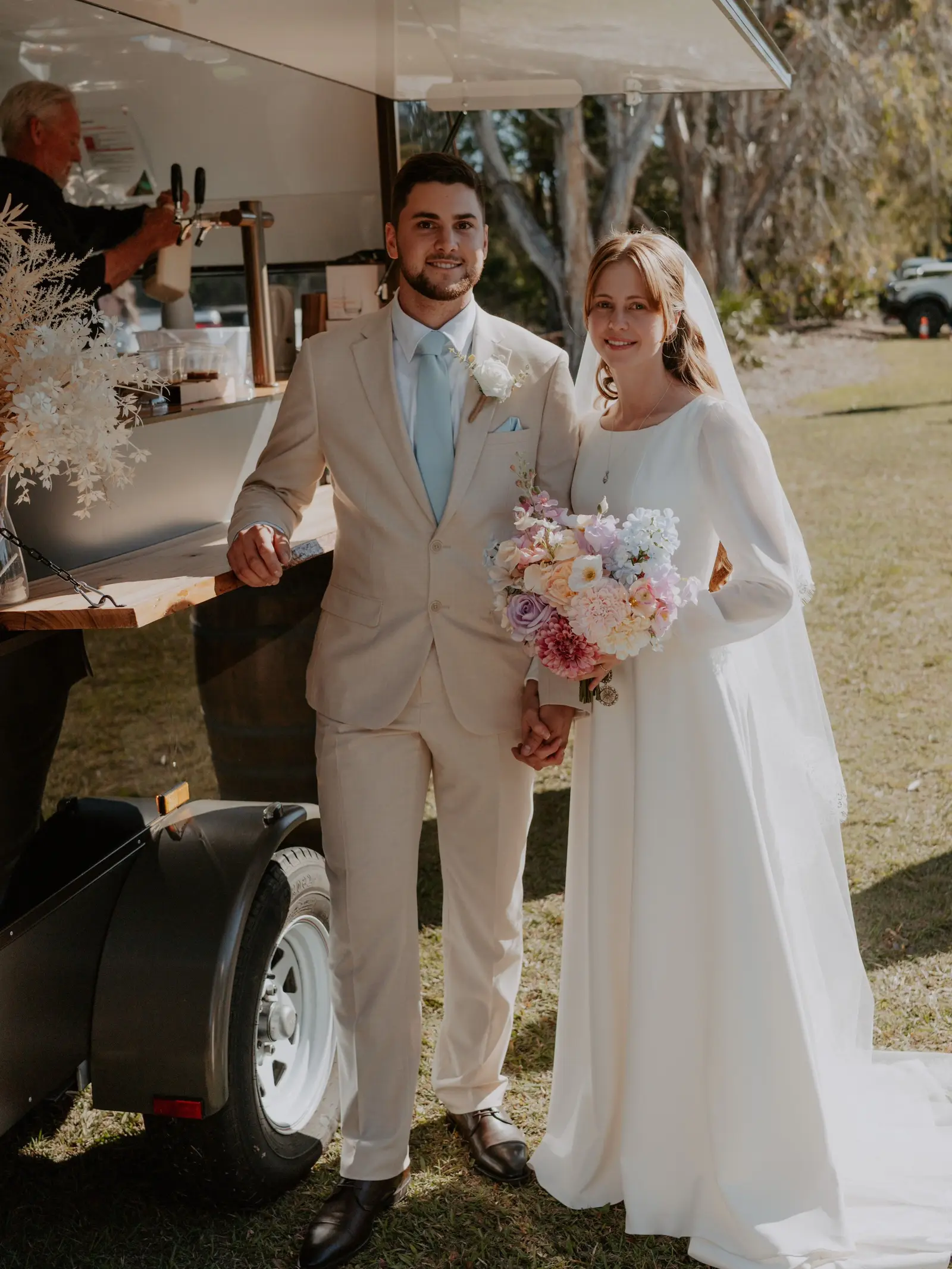 Bride and groom holding hands beside the Whiskey N Wine mobile bar van at a sunny outdoor wedding